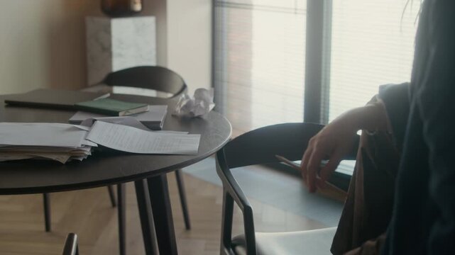 Unrecognizable male author wearing robe, holding pencil in hand sitting in living room during break time, paper drafts, tea mug and laptop visible on round table