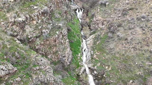 4K aerial drone footage of cascading waterfall in rocky mountain canyon, flowing stream through natural landscape.