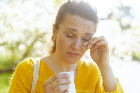 A middle-aged woman expresses discomfort while rubbing her eyes in a bright spring setting. Her struggle with tree pollen allergies is highlighted against a background of green foliage.