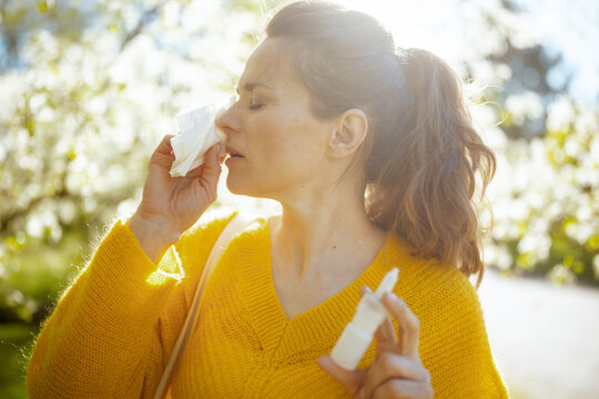 Backlit by warm spring sunlight, a woman in a yellow sweater manages hay fever symptoms with a tissue and nasal spray amidst flowering trees.
