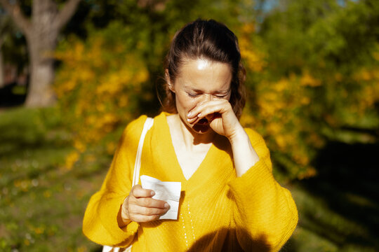 Close-up of a woman in a yellow sweater with her eyes closed in discomfort, holding a tissue while suffering from seasonal allergy symptoms in a blooming spring park.