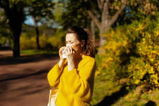 A woman in a yellow sweater uses a tissue while sneezing in a sunlit park, capturing the discomfort of seasonal pollen allergies during the spring bloom.