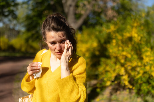 A woman in a yellow sweater rubs her irritated eyes while holding a tissue, depicting common hay fever and pollen allergy symptoms in a spring park.