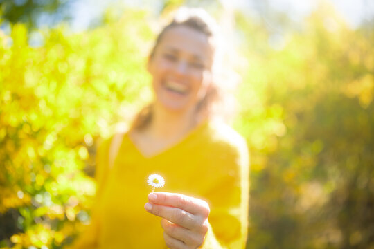 A soft-focus portrait of a woman holding a daisy, with sun-drenched golden foliage in the background. A beautiful representation of mindfulness and spring happiness.