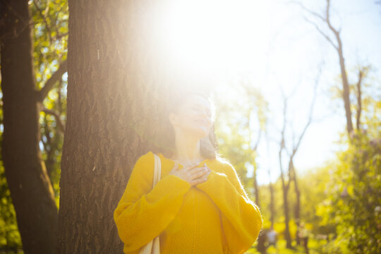 A woman stands with eyes closed and hands on her chest, leaning against a tree, capturing a peaceful moment of deep mindfulness and self-love in spring.