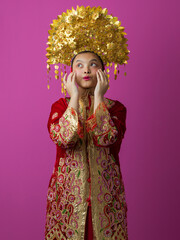 Fototapeta premium Young Indonesian Asian Woman in Ornate Gold Headdress and Embroidered Red Traditional Costume on Pink Background