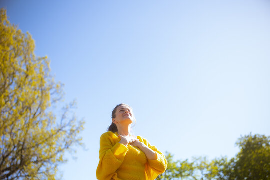 Soft sunlight shines on a smiling woman holding her heart, capturing a beautiful, intimate moment of emotional wellness and self-care within a blooming spring landscape.