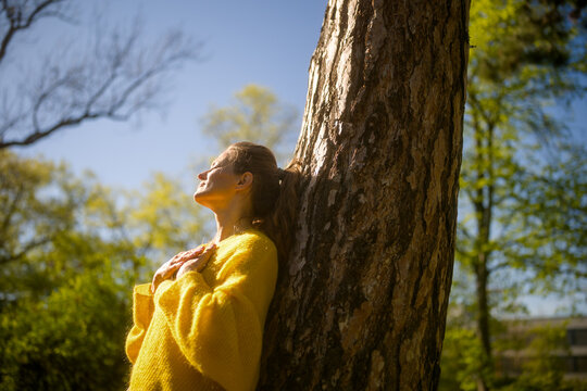 A woman in a yellow sweater leaning against a tree, eyes closed, breathing deeply to find mental stillness, serenity, and connection with nature in the spring sun.