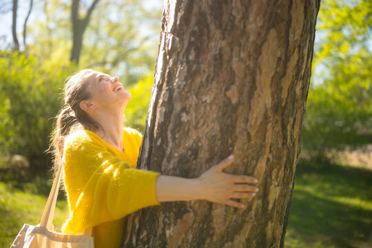 Surrounded by trees, a woman in a yellow sweater embraces a forest giant, her smile reflecting the joy and freedom found in eco-conscious outdoor living.