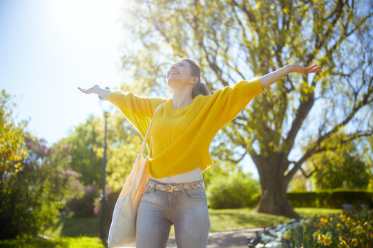 A woman in a yellow sweater with arms outstretched, soaking up the sun in a blooming park, embodying spring joy, freedom, and deep connection with nature.