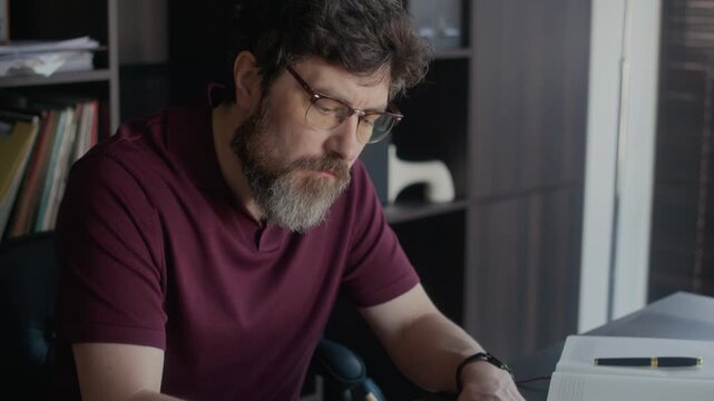 Medium close up of middle aged Caucasian writer proofreading his essay and taking notes while sitting at wooden desk in modern home office