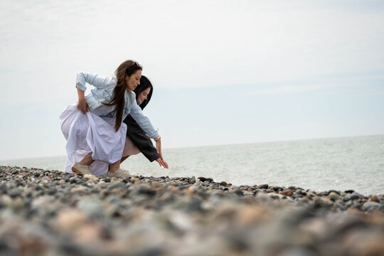 Two young women crouching on a pebble beach by the sea, touching stones and enjoying a quiet coastal moment under an overcast sky.