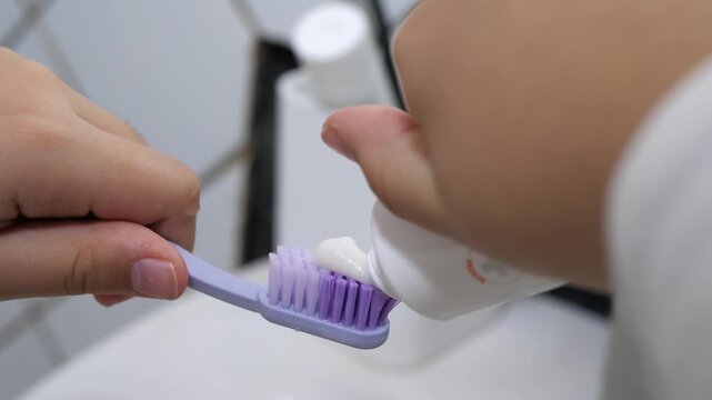 Mother applying toothpaste on a child's purple toothbrush in the bathroom