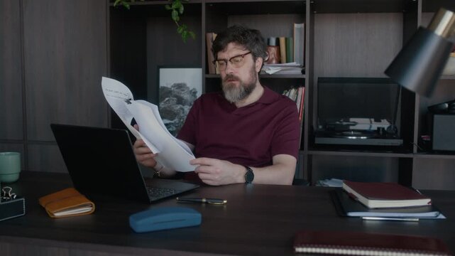 Medium shot of bearded Caucasian male editor reading printed short story draft and writing e-mail to author via laptop at work in modern office