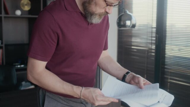 Medium shot of Caucasian male author wearing eyeglasses walking along home office proofreading new short story draft