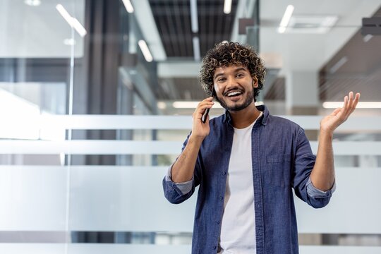 Portrait of a smiling young Indian man standing in the office, talking on the phone and looking at the camera