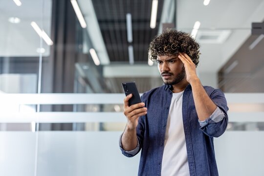 Worried young Indian man standing in office, reading message on phone and holding head in fear