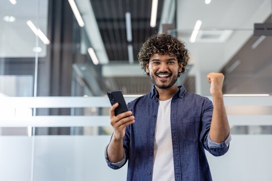 Portrait of a young happy Indian man holding a phone in one hand and showing a victory gesture to the camera with the other hand
