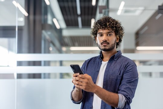 Portrait of a serious young Indian man standing in the office, holding a phone and looking confidently at the camera