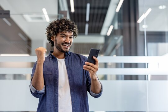 Happy young Indian man standing in the office, looking at the phone screen and rejoicing, showing a victory gesture with his hand