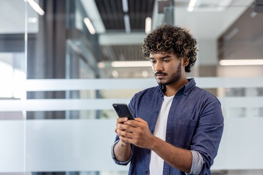 Close-up photo of a serious young Indian man standing in an office building and using his mobile phone