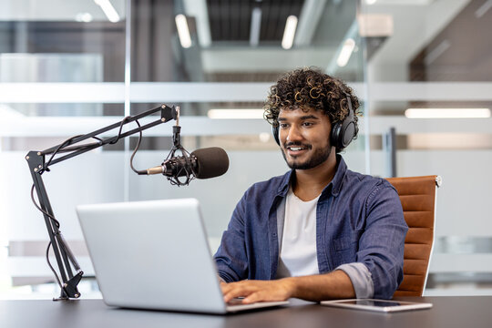 A young Indian man wearing headphones sits at a desk in an office in front of a microphone and works on a laptop