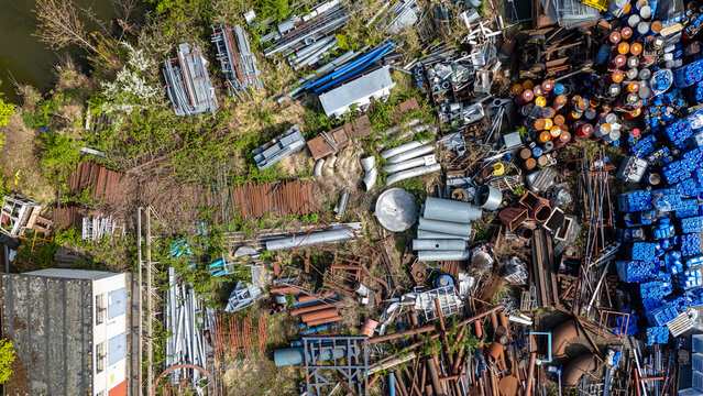 Top-down aerial view of an industrial scrap yard and storage area featuring piles of metal pipes, steel profiles, blue plastic barrels, and rusty construction elements scattered on grassy ground.