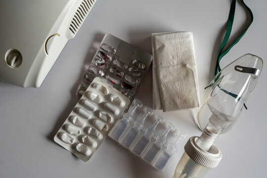Inhalation set featuring a white compressor nebulizer, transparent oxygen mask, saline solution ampoules, medicine blister packs, and paper tissues arranged on a light surface, top-down view, natural 