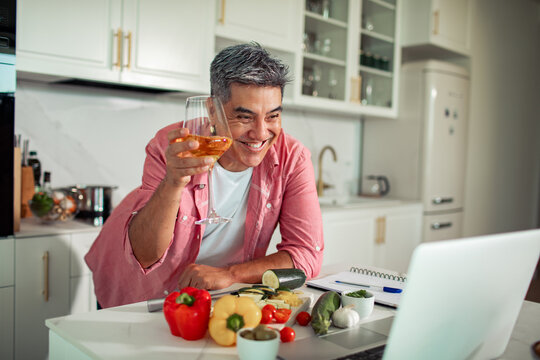 Smiling man toasting with wine on a video call while cooking in a home kitchen