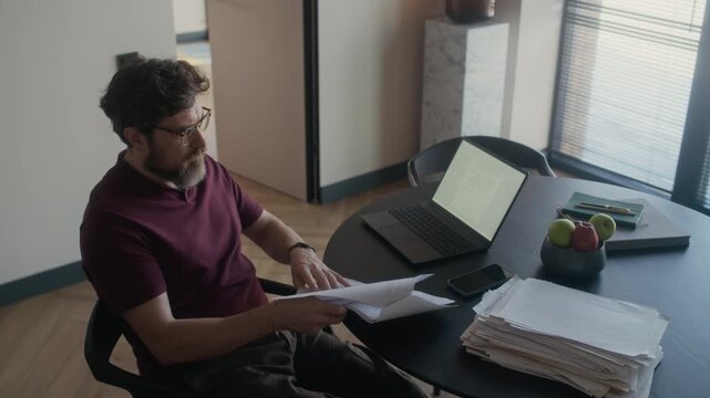High angle view of mature adult Caucasian male author reading printed novel draft at table in living room