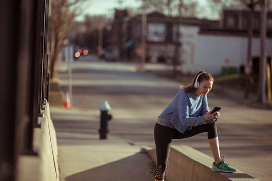 Young woman runner stretching and checking phone on city street