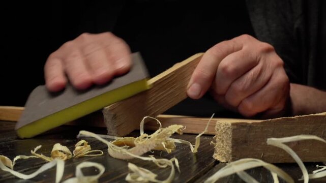 Hands of a craftsman using a sanding block on a wooden piece, surrounded by wood shavings on a dark work surface in a workshop setting
