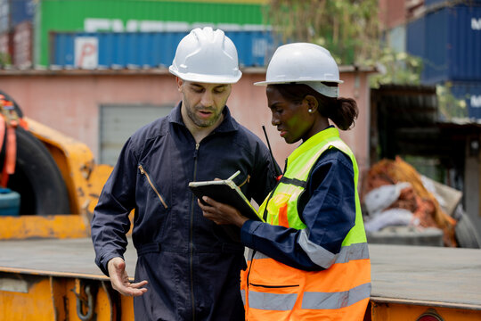 Two industrial engineers workers in hard hats review plans on a tablet at a construction site, coordinating tasks amid containers and heavy equipment.