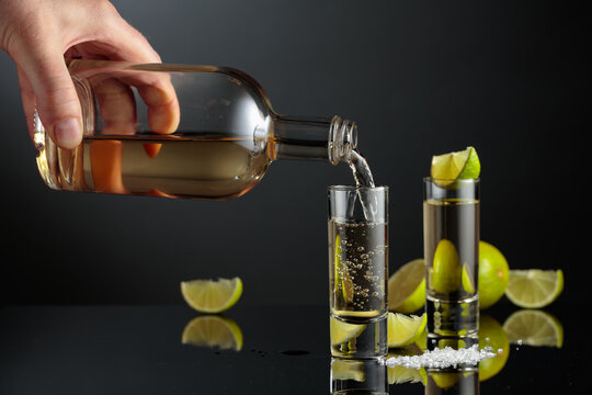 Tequila shots with salt and lime slices on a black reflective background.