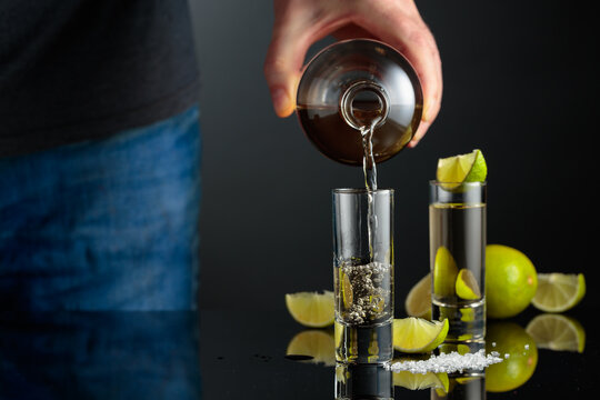 Tequila shots with salt and lime slices on a black reflective background.