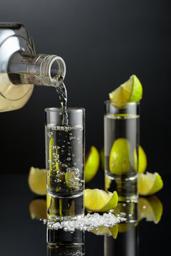 Tequila shots with salt and lime slices on a black reflective background.