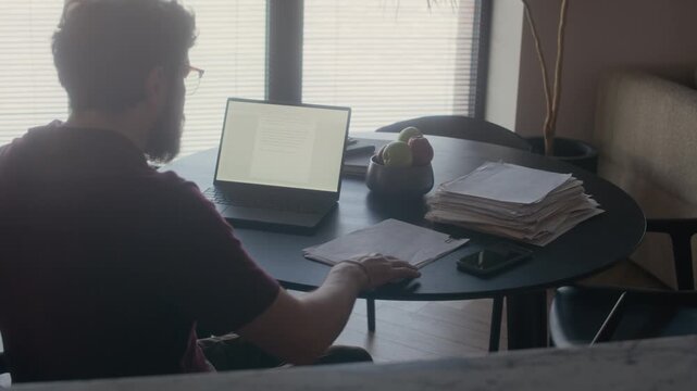 Backlight high angle shot of male Caucasian writer taking seat at table in living room and reading printed novel drafts, perfect for modern author lifestyle content