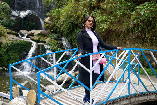 Stylish young woman in a black coat posing on a blue bridge with a scenic waterfall background at Rock Garden Darjeeling