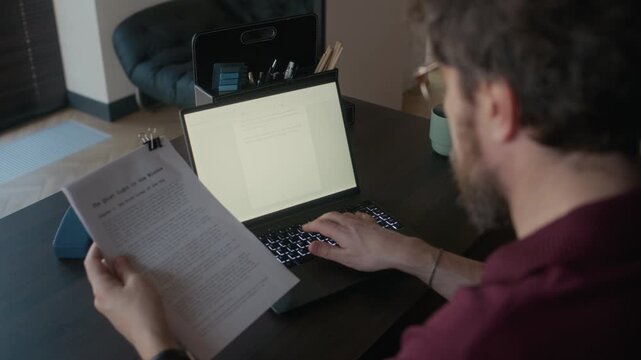 High angle over shoulder shot of bearded male writer looking at printed short story draft and typing final version on laptop
