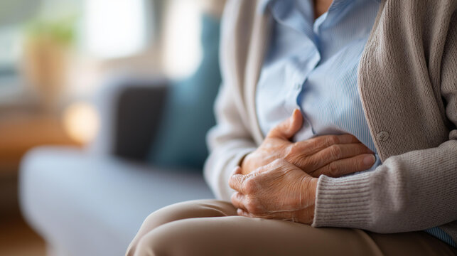 Close-up of elderly person's hands clutching their stomach in apparent pain while sitting on a couch in a blurred living room background. Defocused living room. Elderly hands