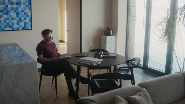 Wide shot of middle aged Caucasian male writer working on novel at table in living room, reading printed drafts and thinking over plot