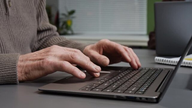 Side view close up shot of wrinkled hands slowly typing on laptop keyboard and using touchpad while unrecognizable senior man browsing internet at desk in class, copy space