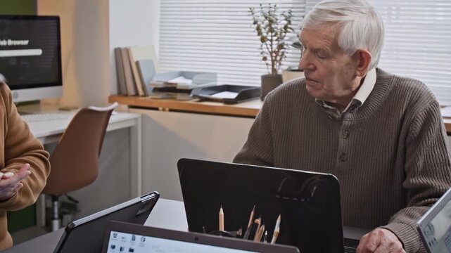 Tilt up shot of white haired senior man using laptop during computer literacy class for seniors and listening to smiling male teacher talking to group at desk in classroom