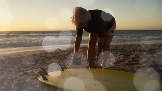 Senior woman bending over yellow board on shore adjusting leash for surfing with flare over hands