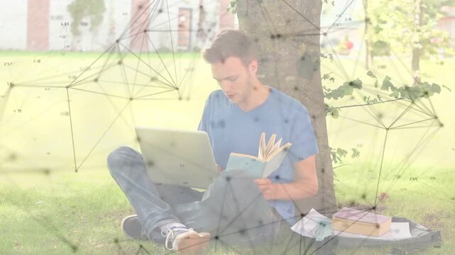 Male student sitting under tree, flipping book, typing laptop, creating network overlays for study