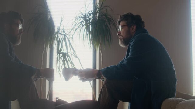 Side view of mature adult Caucasian male writer wearing robe sitting on couch back reading novel draft, his reflection visible on counter surface