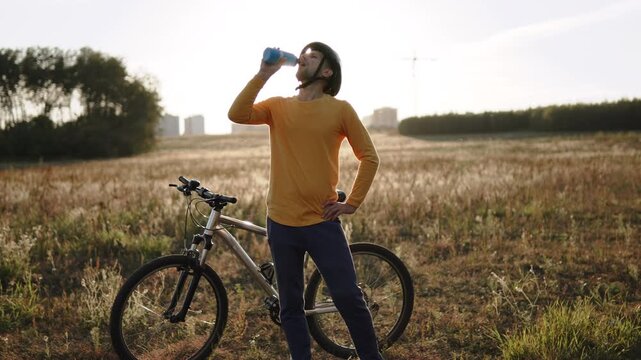 Cyclist drinking water from sports bottle during stop in field after cycling at sunset. Countryside walk of man living active lifestyle, bike riding.