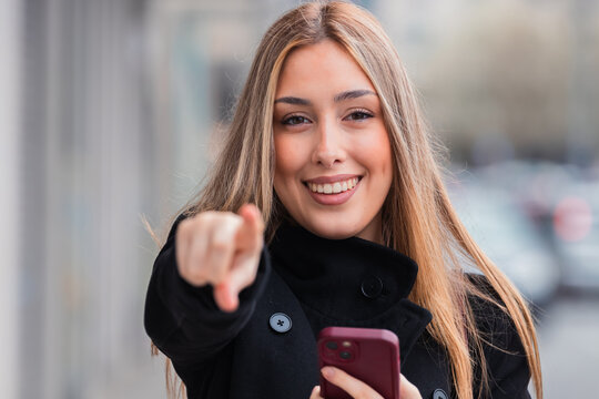 smiling young woman pointing at camera with mobile phone