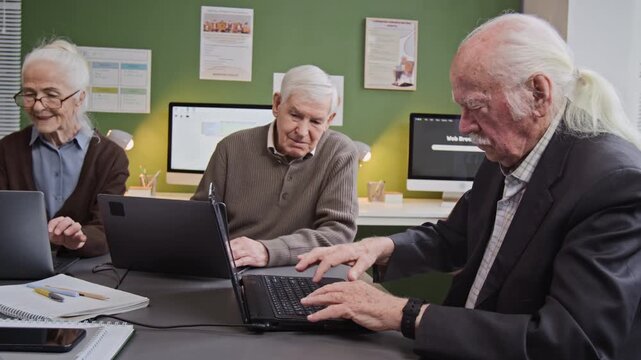 Side view shot of white haired senior man carefully typing on laptop keyboard while doing tasks in computer literacy class sitting in at desk with group of adult students, copy space