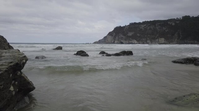 Static shot of rocky shoreline with waves lapping and tide ebbing against cliff and beach under overcast cloudy sky